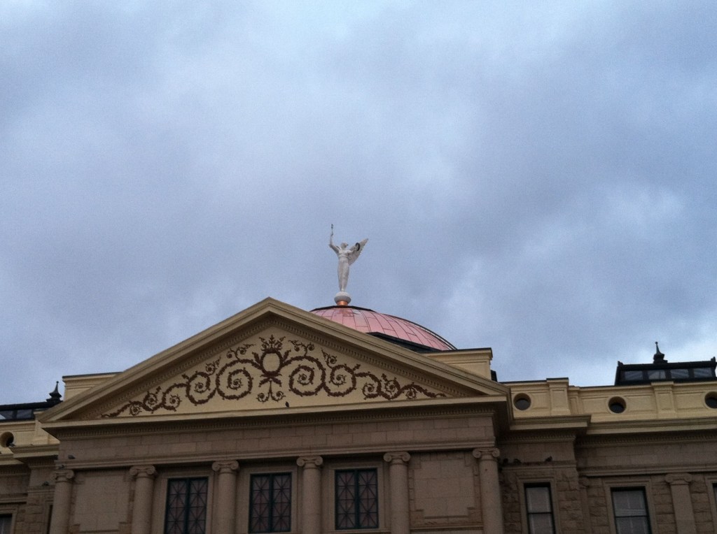 Marble statue of winged victory atop Arizona State Capitol seen against cloudy sky.