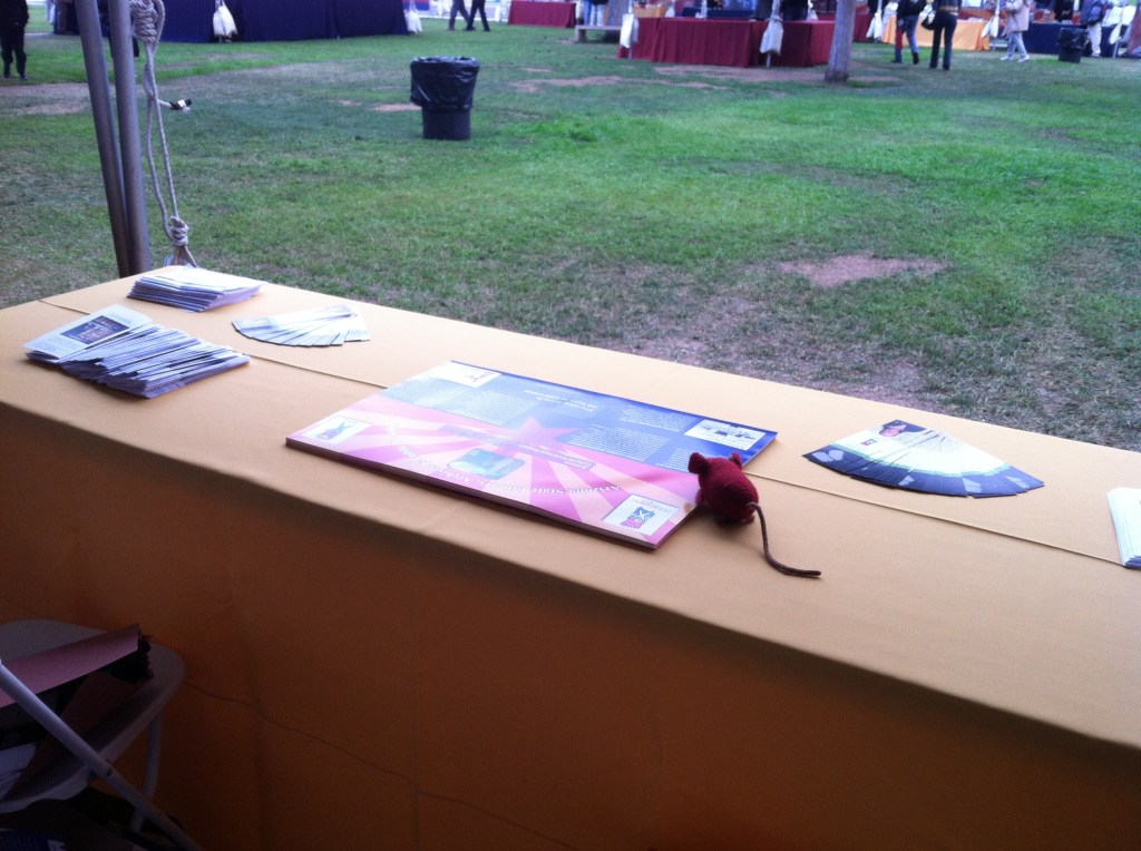 Mousie at the Table Knitted red mouse on a table of posters and trifolds.