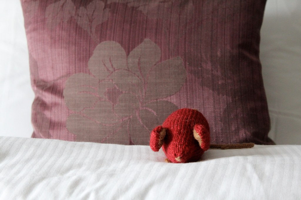 Red knitted mouse on a white duvet with a purple brocade pillow.