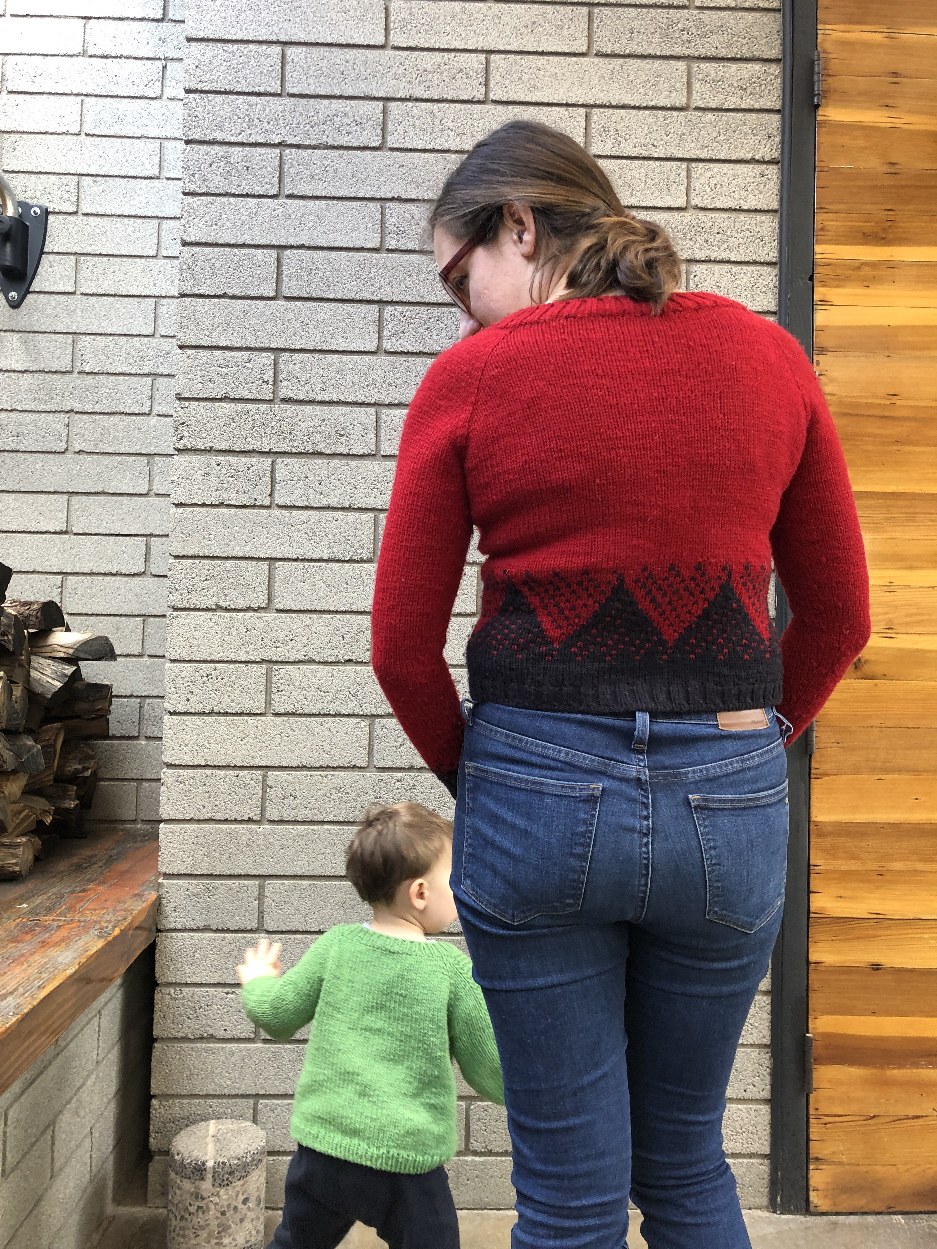 Back view of a woman wearing a red handknitted sweater with a geometric black mountain pattern with a child running behind her