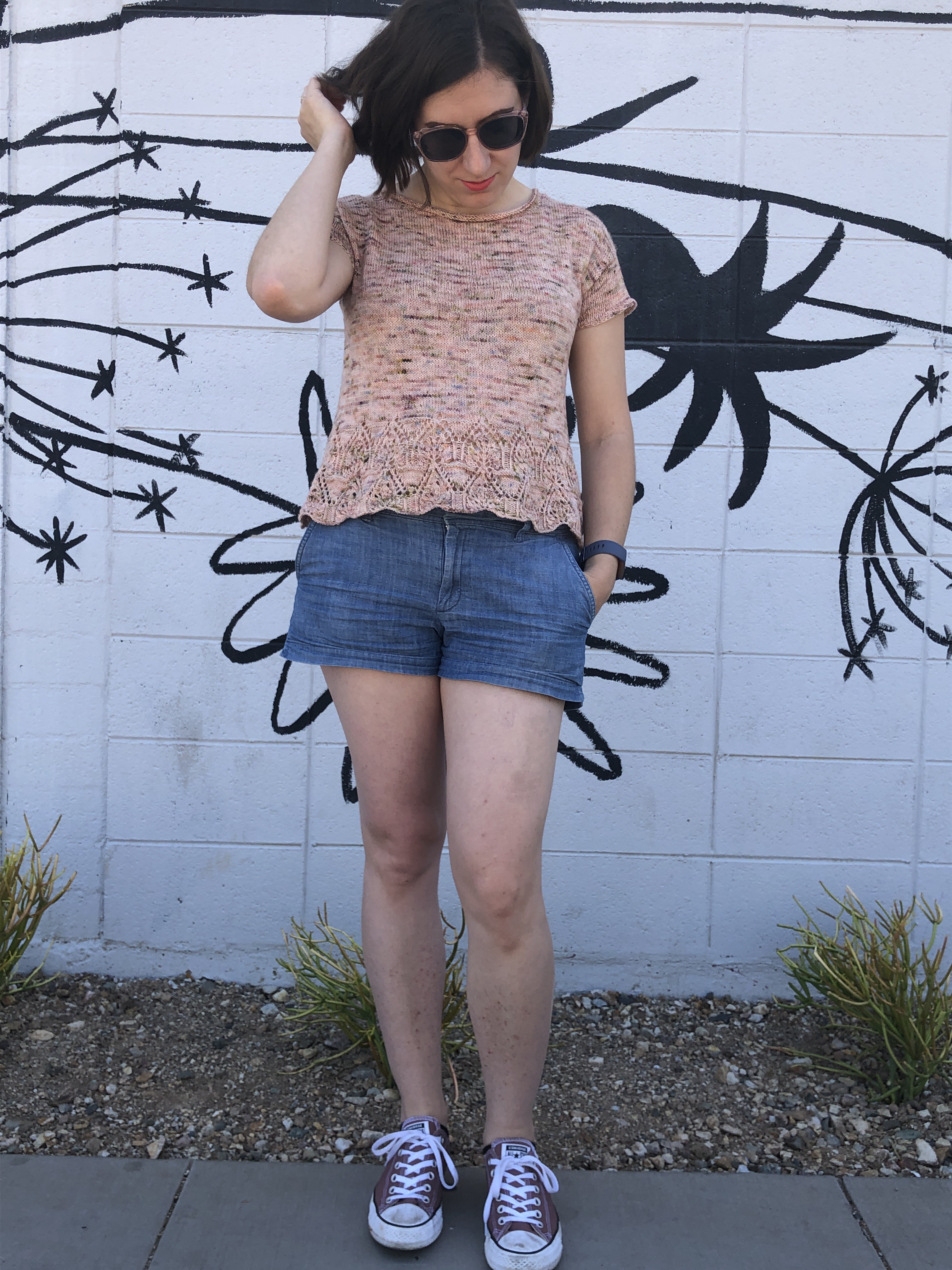 Woman in a pink knitted Tegna top looking away from the camera and adjusting her hair in front of a white wall painted with the outlines of wildflowers