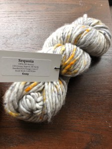 Twisted skein of bulky Baah! Sequoia yarn in Cozy, a gold-speckled gray, on a dark wood surface